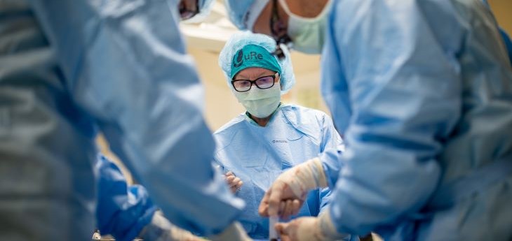 Three people in a surgery operating room are wearing blue surgical gowns, scrub caps, and white masks and are bent over intently over the table.