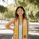 Young woman with shoulder-length brunette hair stands in the street with trees behind her. She is wearing a yellow graduation stole and a white dress.