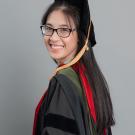 Young woman with glasses wearing doctorate regalia turns and smiles at the camera. She has long black hair and is standing in front of a gray background.