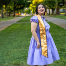 A young woman with short black hair smiles with her chin up. She is wearing a lavender dress and a UC Davis graduation stole.