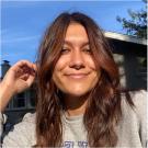 A young woman smiles at the camera and touches her long brunette hair. She is wearing a grey t shirt.