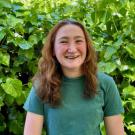 A young woman with wavy brown hair smiles in front of greenery. She is wearing a green t-shirt