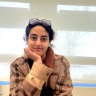 A young woman rests her chin on her hand and smiles at the camera. She has curly black hair that is tied back and glasses on top of her head.