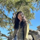 Yougn woman with long black hair smiles with a blue sky and a tree in the background. She is wearing a light green blazer.